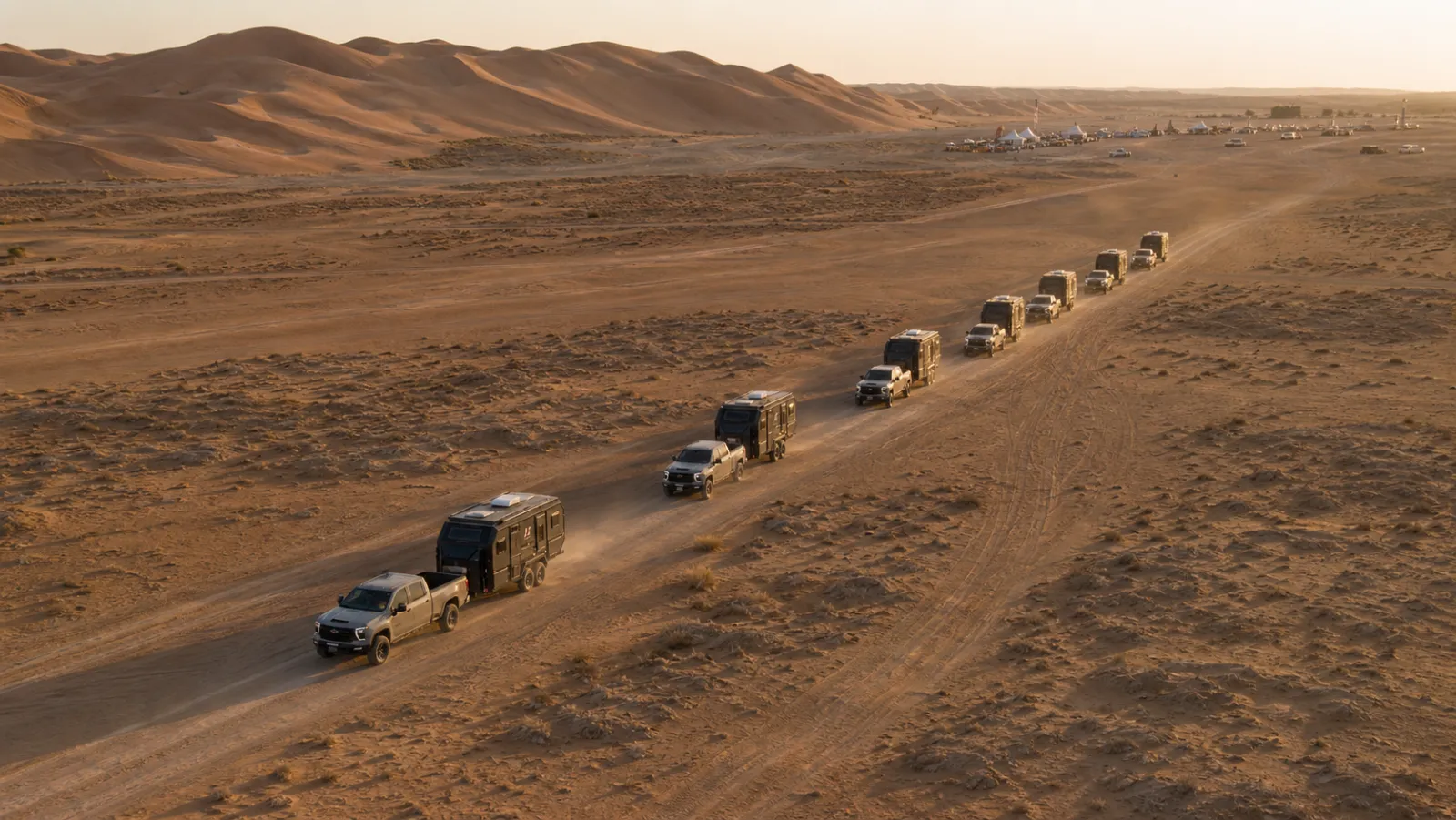 Six pickups towing black expedition caravans in single file along a desert track at dawn, an event camp visible far ahead